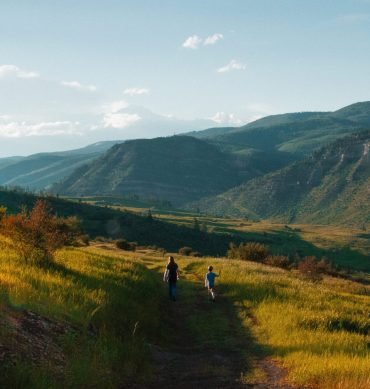 A beautiful shot of a kid and a male walking on a pathway in the middle of grassy fields with forested mountains in the distance